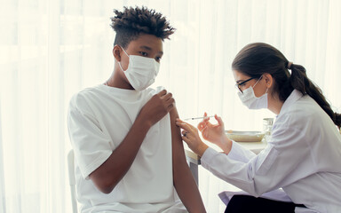 African teen man wearing face mask, getting vaccination to protect or prevent virus, getting scary vaccine injection while female caucasian doctor preparing syringe to vaccinate patient at hospital. - Powered by Adobe