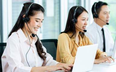 Focus Asian beautiful attractive female call center cheerful smiling with headsets, sitting, typing, monitoring with colleagues in office or agency at computer, giving service to customers.