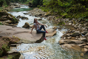 Happy smiling girl in glasses, white t-shirt, blue jeans and red sneakers sitting near mountain river. Young woman enjoys fresh air in forest resting on a rock above a waterfall. lonely traveler.