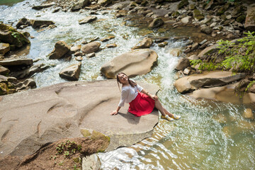 Happy smiling girl in a white blouse, red skirt and yellow sneakers sitting near mountain river. Young woman enjoys fresh air in forest resting on a rock above a waterfall. lonely travel