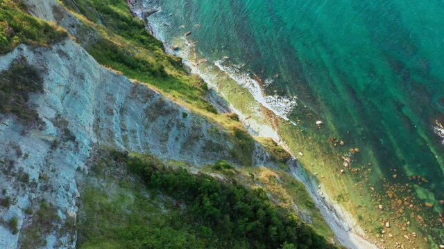 Aerial view of rocky coastline, down blue sea, deep white cliffs. White rocks, trees. Sea waves in blue seaside in the background, blue sky in a summer day in Conero, Italy