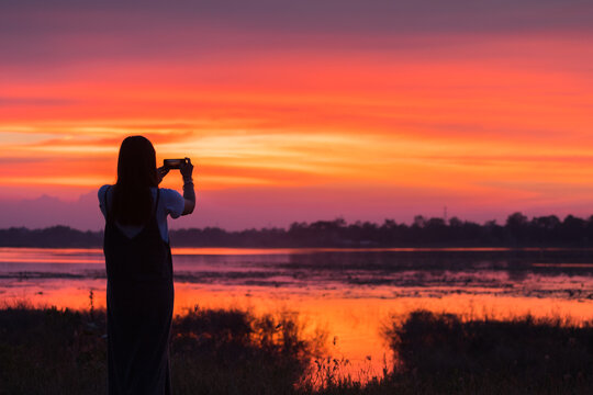 silhouette of a woman holding a cell phone taking pictures outside during sunrise or sunset on the lake. 