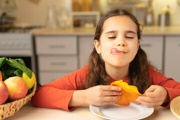 Cute kid girl is happy to eat mangoes. Schoolgirl joy tasty eating and yummy a half of mango on table at home in kitchen