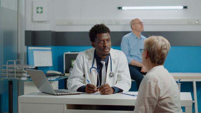 Young Doctor With White Coat And Medical Stethoscope Discussing Healthcare Treatment With Elder Patient In Cabinet . Medic Having Conversation With Woman While Senior Man Sitting On Bed