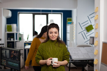 Fototapeta na wymiar Portrait of designer artist smiling at camera holding cup of coffee standing in start up multimedia agency office. Woman working in creativity studio production editing video design with diverse team.