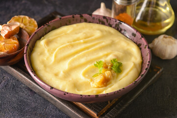 Bowl of tasty mashed potatoes with garlic and ingredients on dark background, closeup