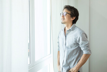 Middle-aged Asian man wear glasses in grey shirt standing by the window at home.