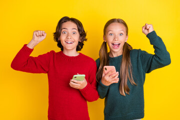 Photo portrait two children keeping mobile phones gesturing like winner isolated vibrant yellow...