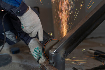 The cut off male hands of a worker use a grinder to clean a weld seam on a metal structure. Bright sparks of hot metal fly.