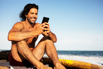 Young man using the phone while sitting on the beach. Handsome man resting after surfing