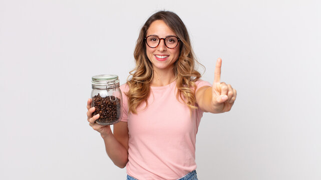 pretty thin woman smiling and looking friendly, showing number one and holding a coffee beans bottle - Powered by Adobe