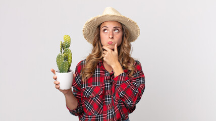 pretty thin woman thinking, feeling doubtful and confused holding a cactus. farmer concept - obrazy, fototapety, plakaty