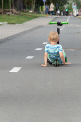 A little boy learns how to control a three-wheeled scooter on a bike path in a city park.
