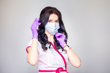 A young woman in medical gloves and a white coat puts on a medical mask.