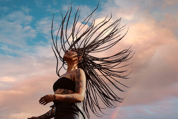 Beautiful young tattooed girl with flying up braids against a blue sky - obrazy, fototapety, plakaty