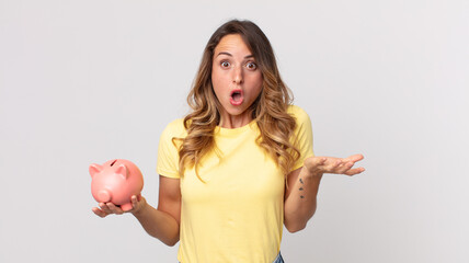 pretty thin woman amazed, shocked and astonished with an unbelievable surprise and holding a piggybank