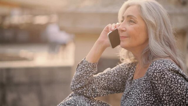 Attractive grey-haired senior woman talking using smartphone with her friends. Happy smiling middle aged woman sat to rest by the cool fountain on a hot day.