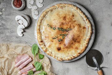Italian calzone pizza with  mozzarella, ham, basil  on marble round tray. Garnished on grey stone backdrop with pizza ingredients