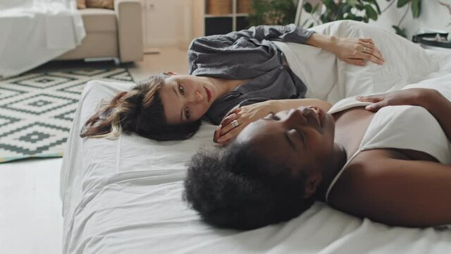Slowmo medium shot of happy Caucasian young woman lying on bed and chatting with her African-American girlfriend while holding hands with her