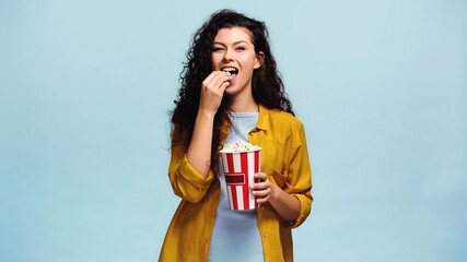 cheerful woman with curly hair eating popcorn isolated on blue
