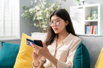 Focused pretty girl of asian beauty is in the living room on the sofa, in hands she holds phone and bank, credit card, the woman is shopping online, payment, enters the number security code