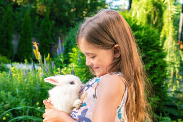 The girl is holding a white fluffy hare on her chest. Happy childhood concept. Pet and kid friendship concept. Rodent rabbit as a pet in the families with children.