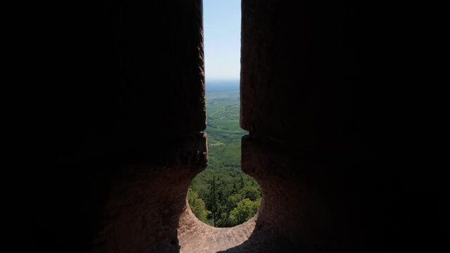Look out to green Alsatian landscape through narrow crosslet loop in wall of old castle. Then camera move back and show dark room with bright embrasure. Forested foothills, vineyards and small village