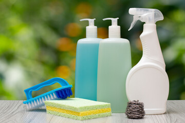 Bottles of dishwashing liquid, glass and tile cleaner, brush and sponges on blurred background.