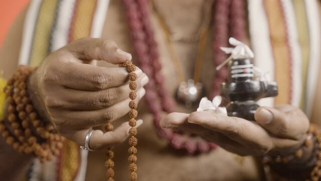 Close up hands of Indian priest meditating or chanting using rudrakshi japa mala with shiva linga in hand during navratri.