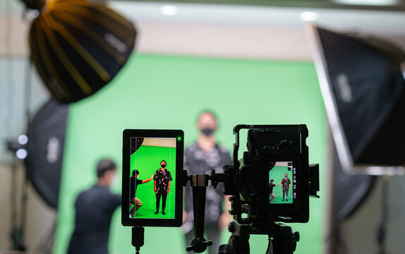 Selective focus of a tablet screen attaching with camera while shooting two Asian men, one standing and the other gesturing on a green screen with a blurred of them in the background in the studio.