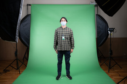 Front view of young Asian man in a gray plaid shirt and navy pants standing on a green screen background with LED softbox lights on tripods illuminate to him, get ready for a shoot in the studio.