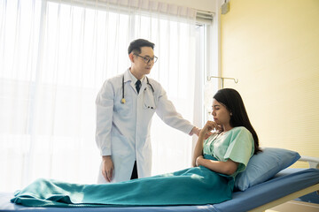 Asian male doctor wearing stethoscope and comforting of unhappy female patient sitting on a hospital bed