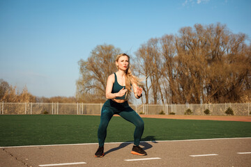 Sporty woman is jumping, doing squats outdoors on the stadium. 