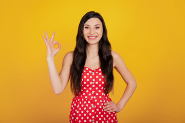 Portrait of positive woman show okey with finger look camera wear dotted red dress on yellow background