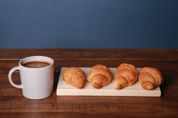 Fresh baked croissants on chopping board, mug of black coffee on brown wooden table. Cup of hot Americano with foam, buns, rolls close up. Food, French breakfast, morning menu, cafe
