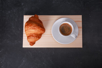Fresh baked croissant on chopping board, wooden pad, white mug of expresso on dark concrete table. Cup of hot coffee, buns, rolls close up. Food, French breakfast, morning menu, cafe concept. Top view