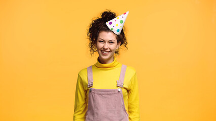 cheerful woman in party cap and yellow turtleneck looking at camera isolated on orange