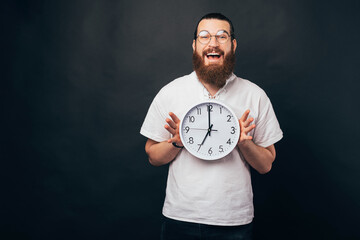 Bearded male hipster is holding a round wall clock while smiling at the camera.