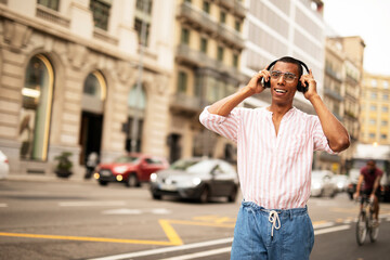 Cheerful guy listening the music with the headphones. Young african man walking through the city