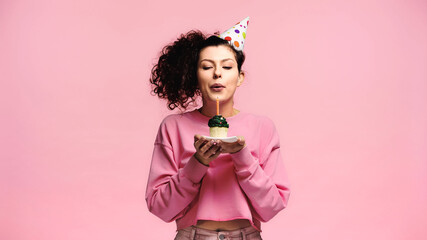 young woman in party cap blowing candle on birthday cupcake isolated on pink