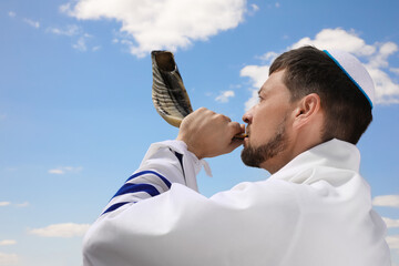 Jewish man in kippah and tallit blowing shofar outdoors. Rosh Hashanah celebration