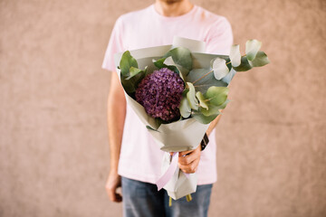 Very nice young man holding simple and beautiful bouquet of fresh eucalyptus and hydrangea in green and purple colors, cropped photo, bouquet close up on the wooden background