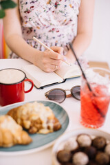 Woman making notes in her notebook, while sitting by the cafe table, drinking coffee, enjoying summer strawberry lemonade, chocolate candy and croissants, with sunglasses and a flower vase standing