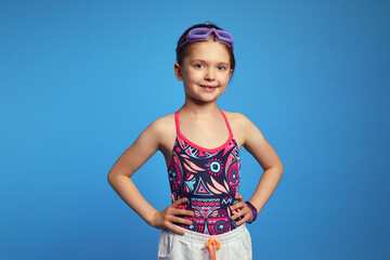 Confident little kid girl holds arms on waist, ready for swim, poses over blue background, wears goggles and swimsuit
