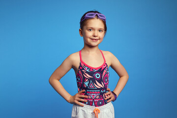 Confident little kid girl holds arms on waist, ready for swim, poses over blue background, wears...