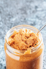 Spoon dipping into a glass jar with crunchy peanut butter. Place for text. Food background.
