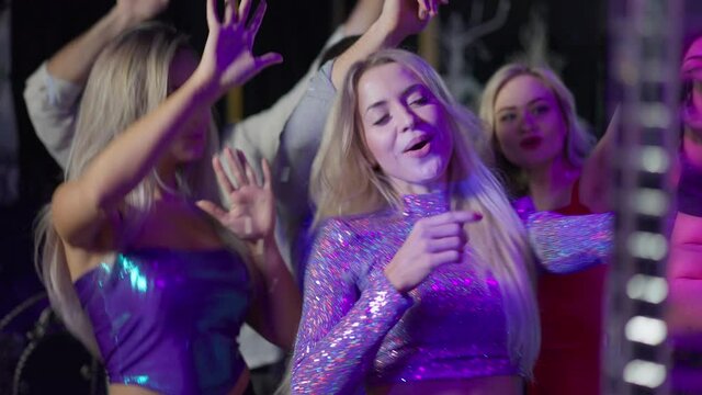 Beautiful happy Caucasian female blogger filming leisure in night club on smartphone dancing with group of friends indoors. Portrait of slim smiling woman streaming online from dance floor