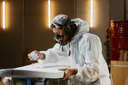 Man in respirator shaping surfboard in workshop - Powered by Adobe