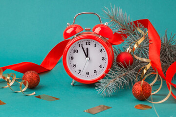 Red alarm clock on a table with red Christmas decor and fir branches on a pastel turquoise...