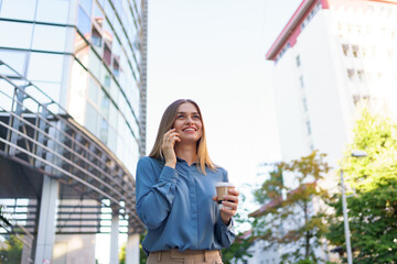 Caucasian business woman speaking by phone holding coffee to go. A successful European woman, talking on the phone, standing on modern office building background